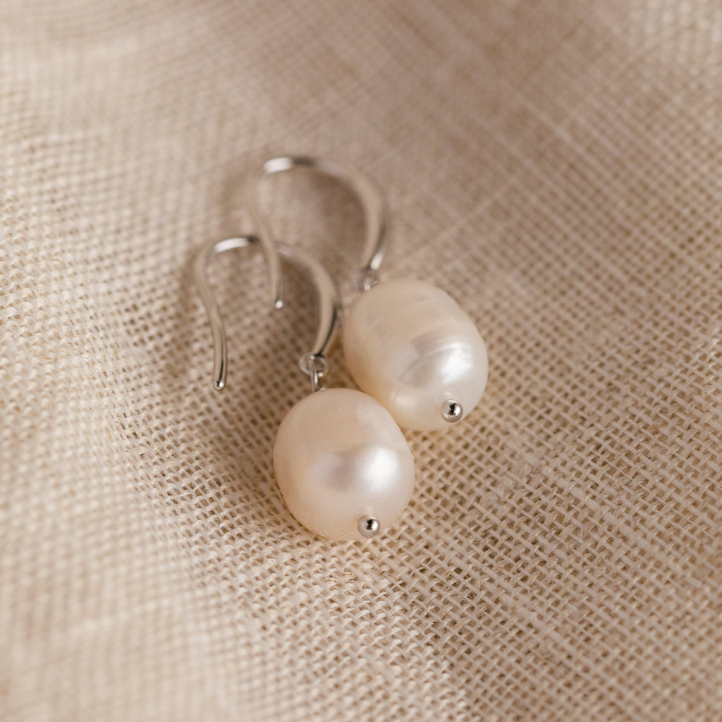 The silver Dangling Pearl Drop Earrings placed on a coarse natural fabric, showcasing their classic shape and luster.
