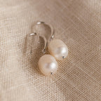 The silver Dangling Pearl Drop Earrings placed on a coarse natural fabric, showcasing their classic shape and luster.
