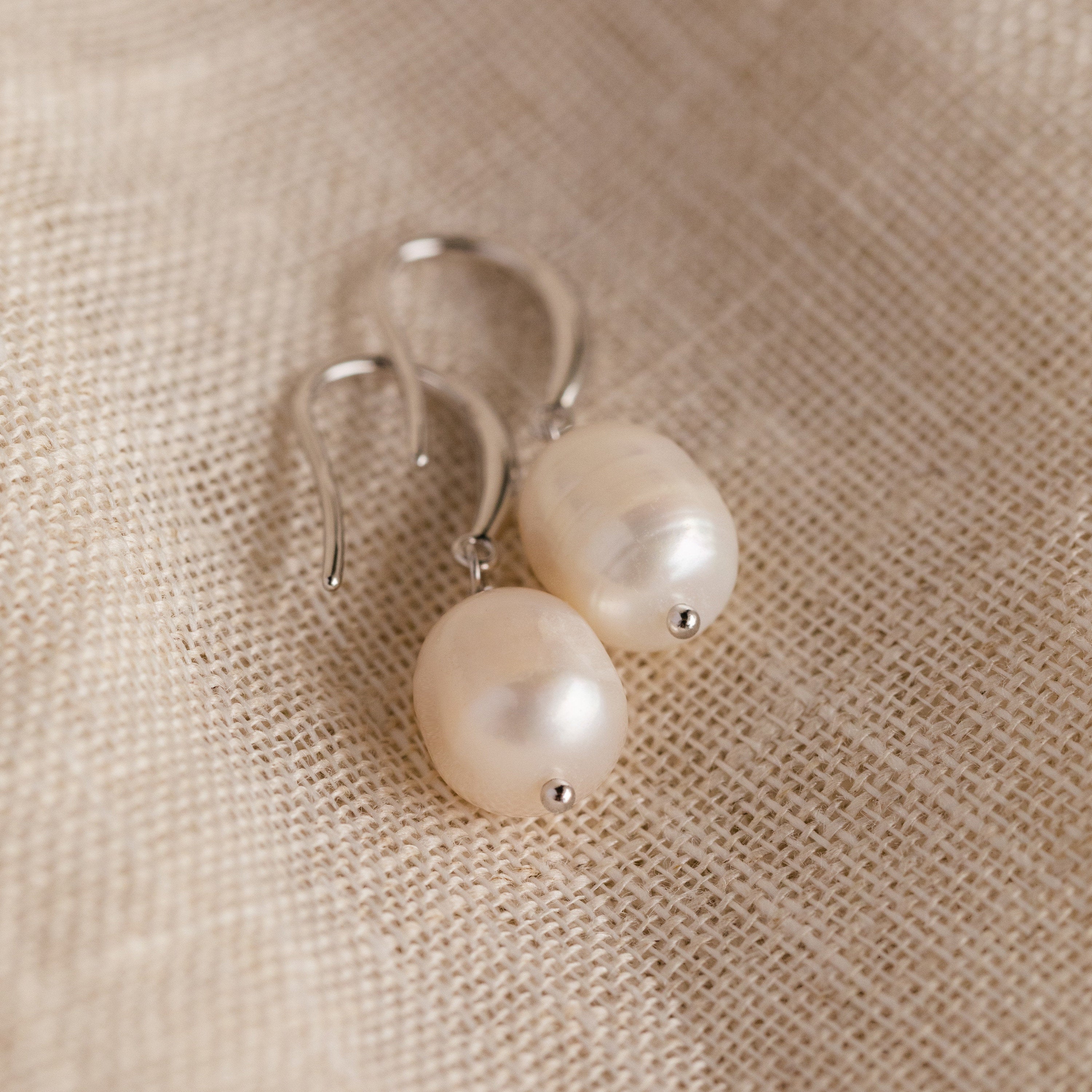 The silver Dangling Pearl Drop Earrings placed on a coarse natural fabric, showcasing their classic shape and luster.