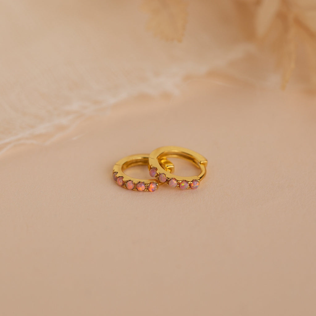 Pink Opal Huggies—gold dainty hoop earrings with small pink gemstones, displayed on a soft beige surface against a delicate fabric background. Elegant, feminine jewelry perfect for any occasion.
