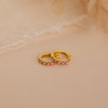 Pink Opal Huggies—gold dainty hoop earrings with small pink gemstones, displayed on a soft beige surface against a delicate fabric background. Elegant, feminine jewelry perfect for any occasion.