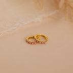 Pink Opal Huggies—gold dainty hoop earrings with small pink gemstones, displayed on a soft beige surface against a delicate fabric background. Elegant, feminine jewelry perfect for any occasion.