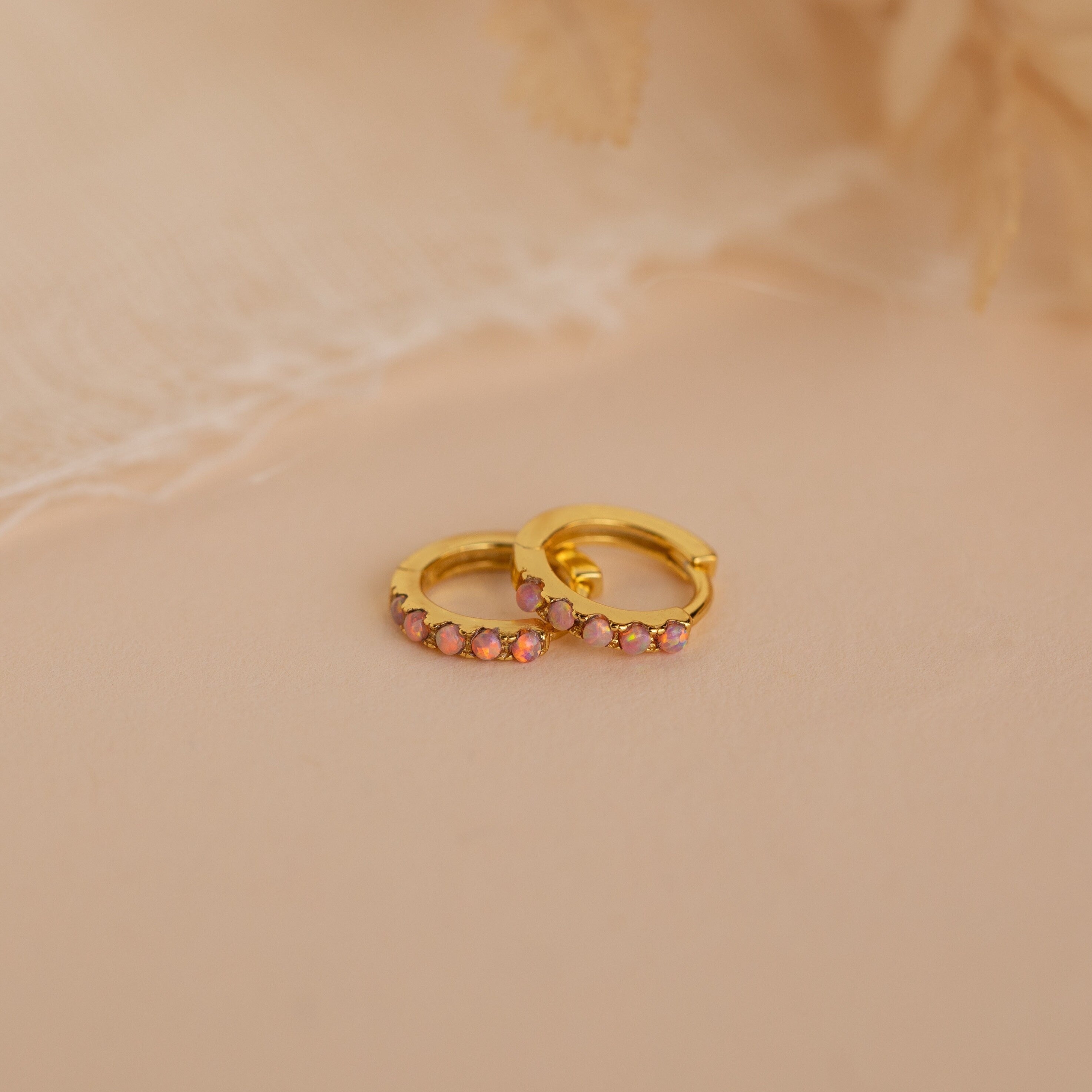 Pink Opal Huggies—gold dainty hoop earrings with small pink gemstones, displayed on a soft beige surface against a delicate fabric background. Elegant, feminine jewelry perfect for any occasion.