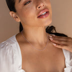 A woman wearing the Pink Station Necklace and a white blouse gently touches her chest against a neutral background.