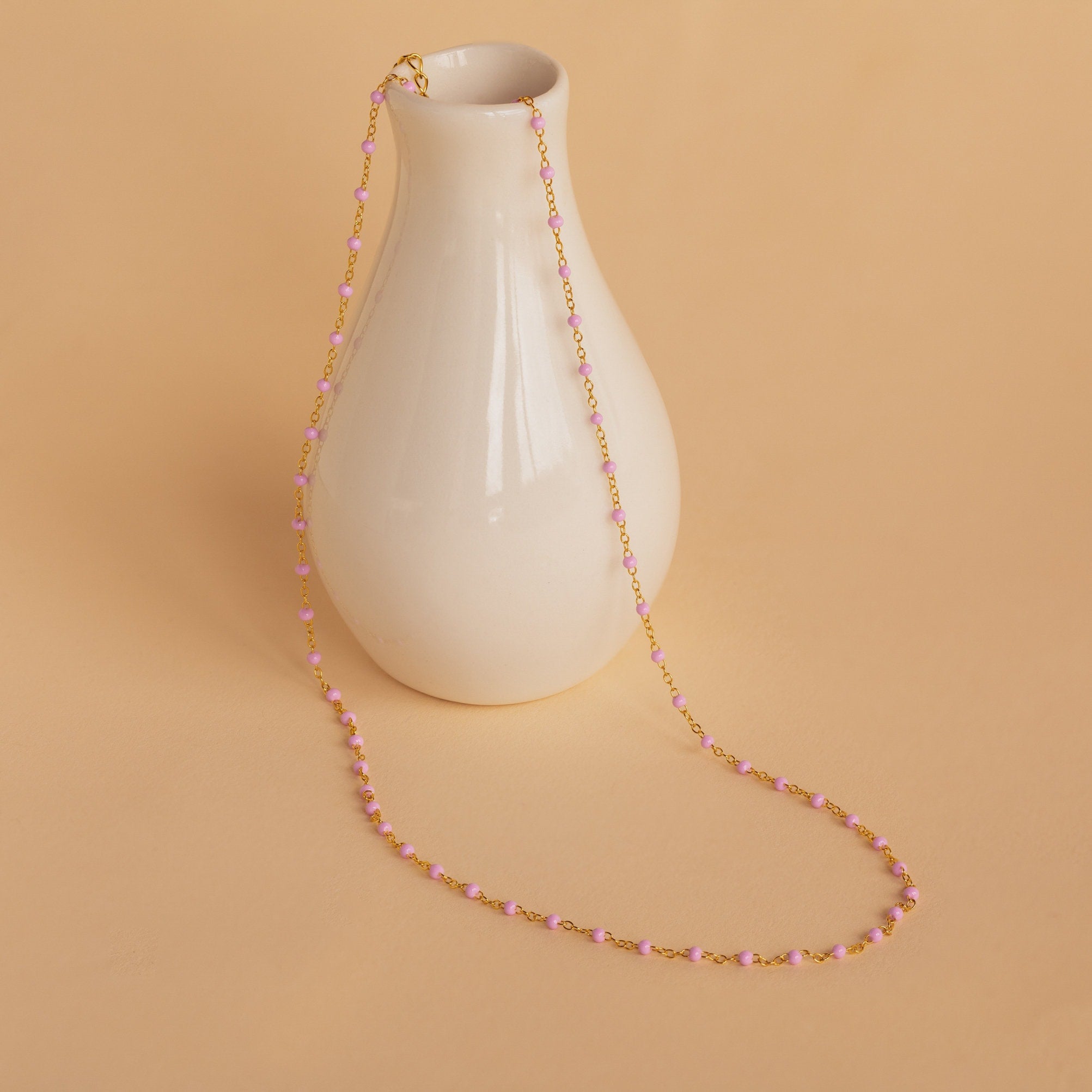 The Pink Station Necklace, featuring gold beads, is displayed draped over a small white vase on a beige background—ideal for layering as a dainty chain.