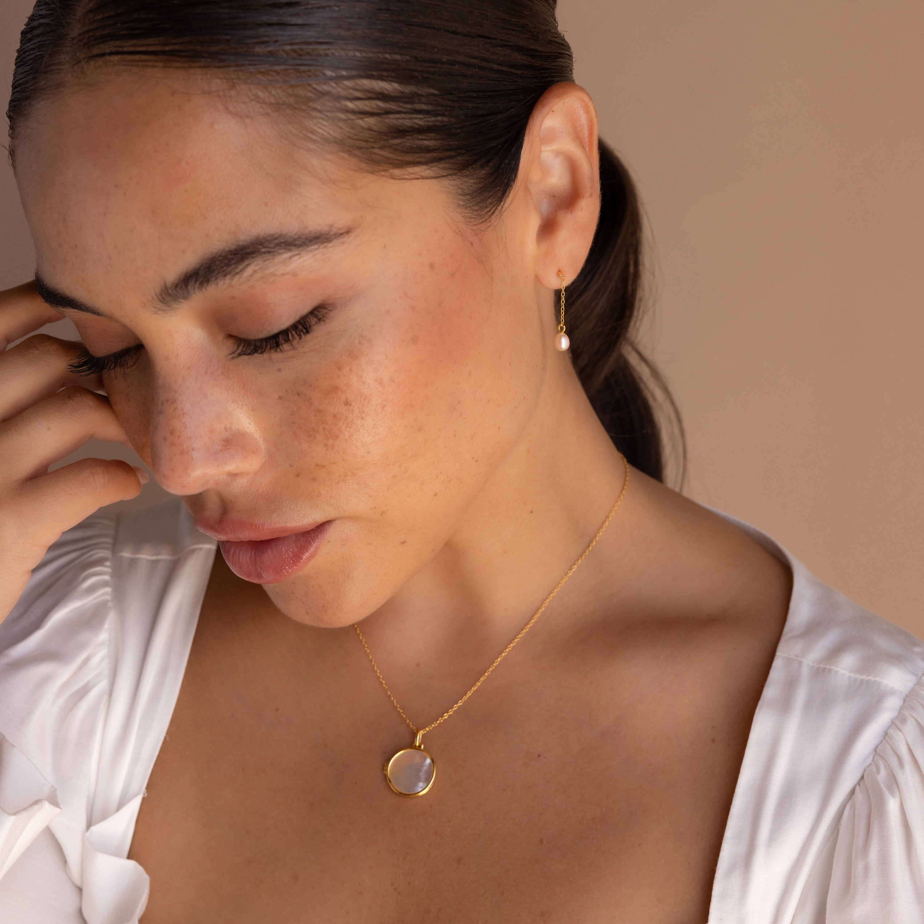 A woman with closed eyes wears Isha Pink Pearl Earrings and a gold necklace with a round pendant, softly illuminated.