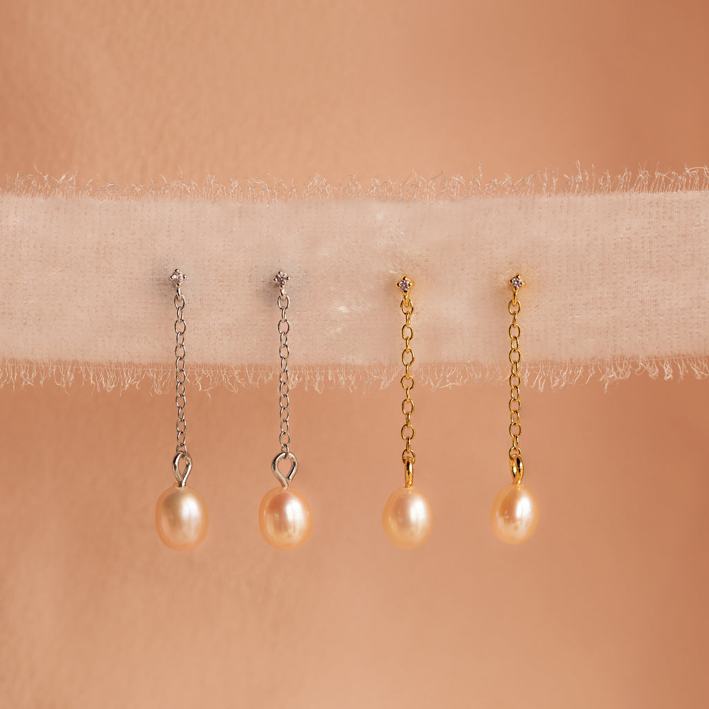 Two pairs of Isha Pink Pearl Earrings—one with silver dangling chains and one with gold—are displayed on soft fabric.