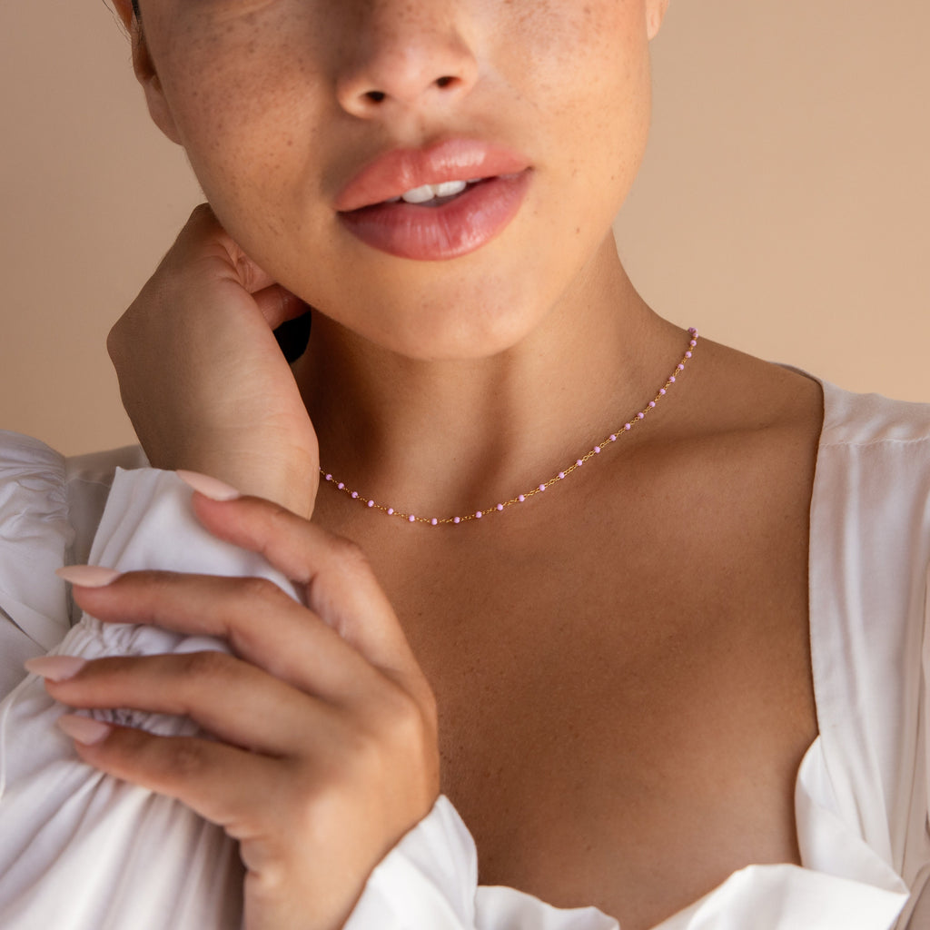 A woman in a white blouse gently touches her neck and collarbone, showcasing the Pink Station Necklace.