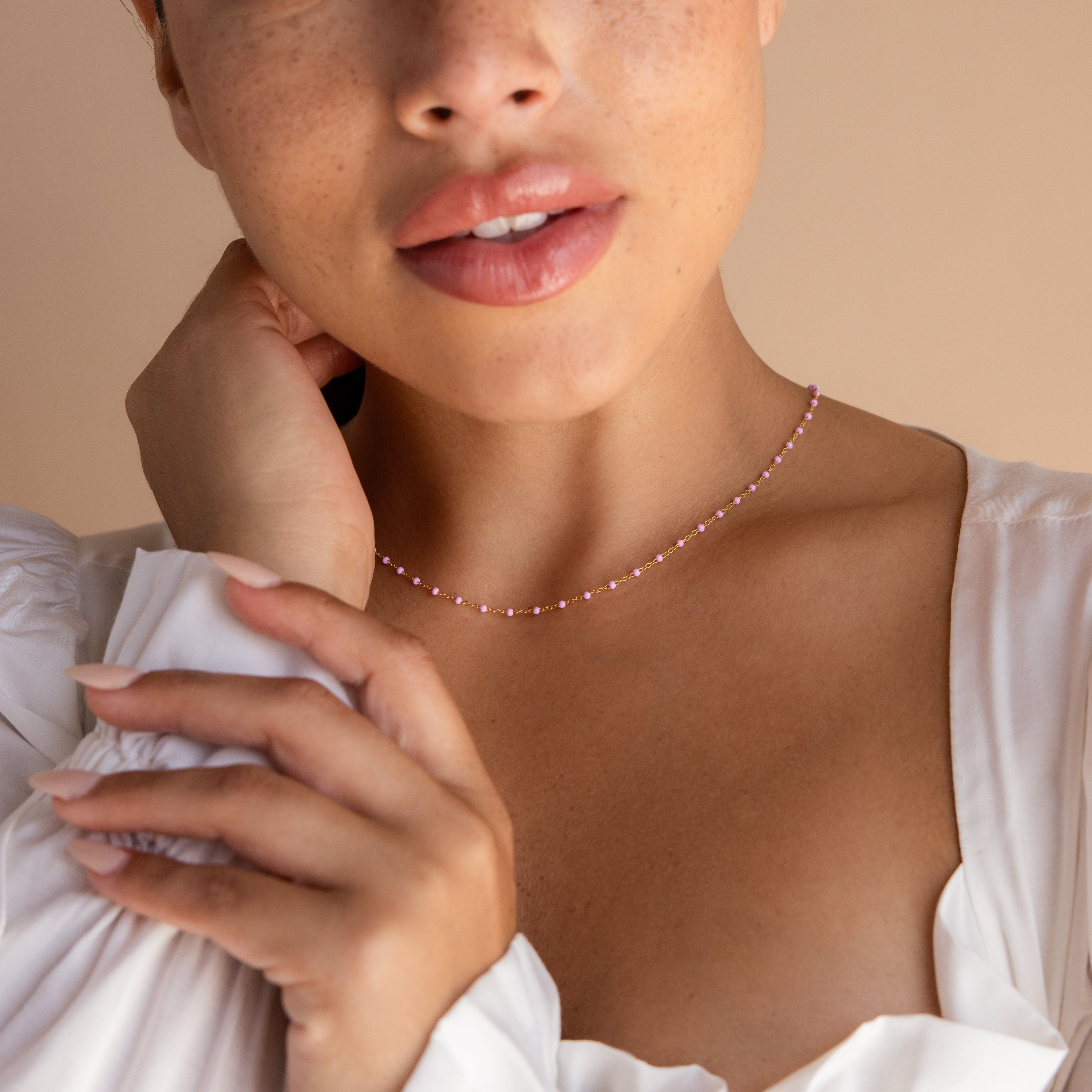 A woman in a white blouse gently touches her neck and collarbone, showcasing the Pink Station Necklace.