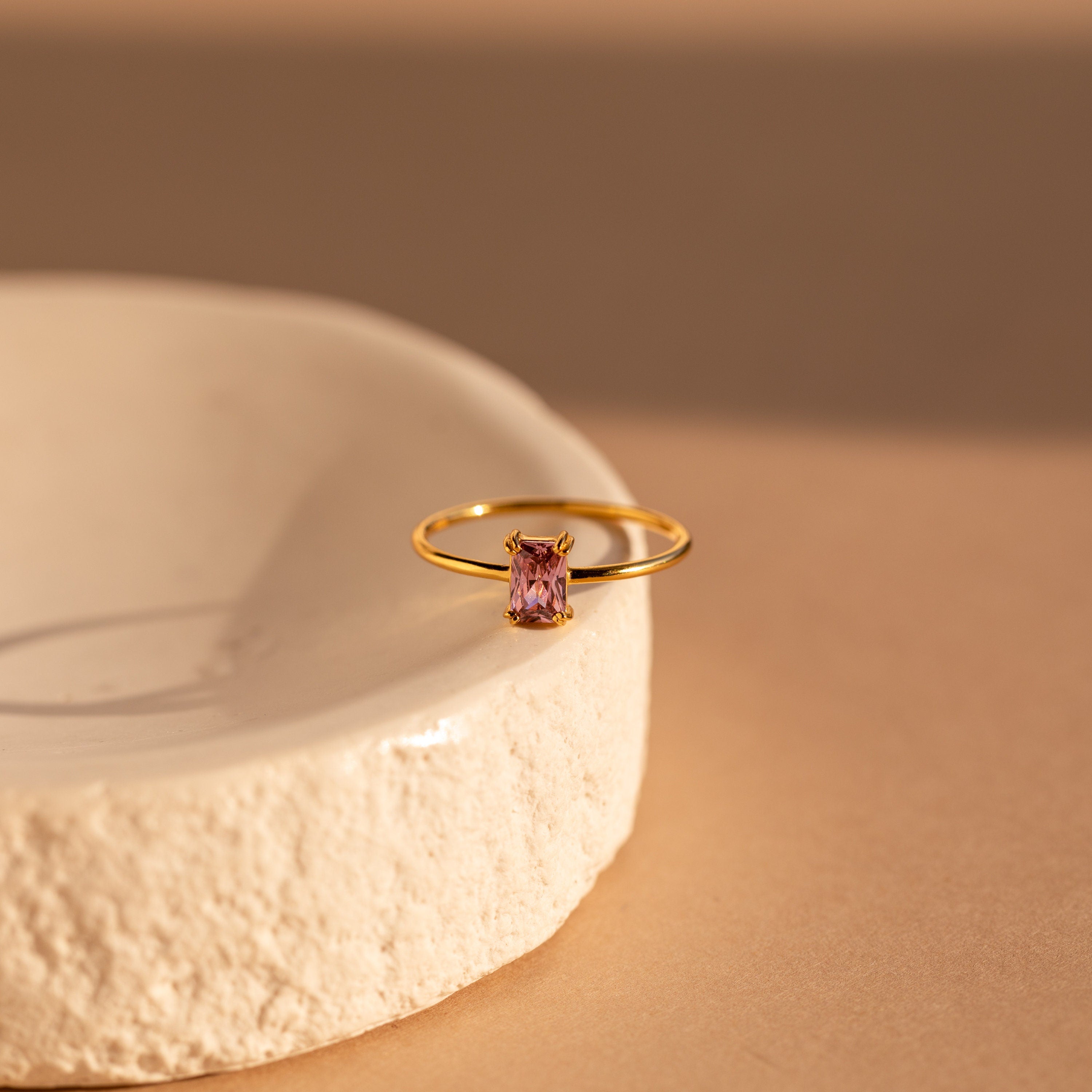 The Blush Pink Emerald Ring, featuring a rectangular gemstone, sits on the rim of a textured white ceramic dish.