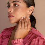 A woman in a blush pink blouse with a sleek ponytail shows off the Blush Pink Emerald Ring, a dainty stackable piece, with her hand near her face.