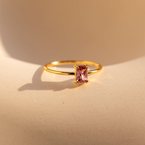 The Blush Pink Emerald Ring features a stackable gold band with a rectangular blush pink gemstone, elegantly casting a shadow on a beige surface.