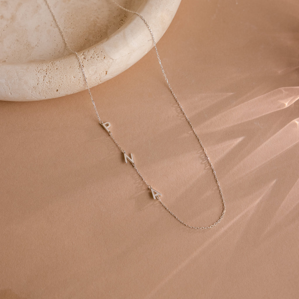 The Sideways Initial Necklace, displayed on a beige surface next to a round stone dish, features gold PNA letters—a unique and thoughtful gift for Christmas.