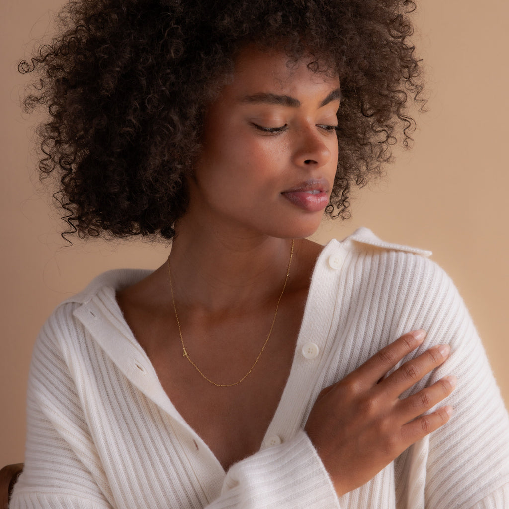 Woman with curly hair wearing a white cardigan and the Venice Sideways Initial Necklace, looking down with a soft expression.