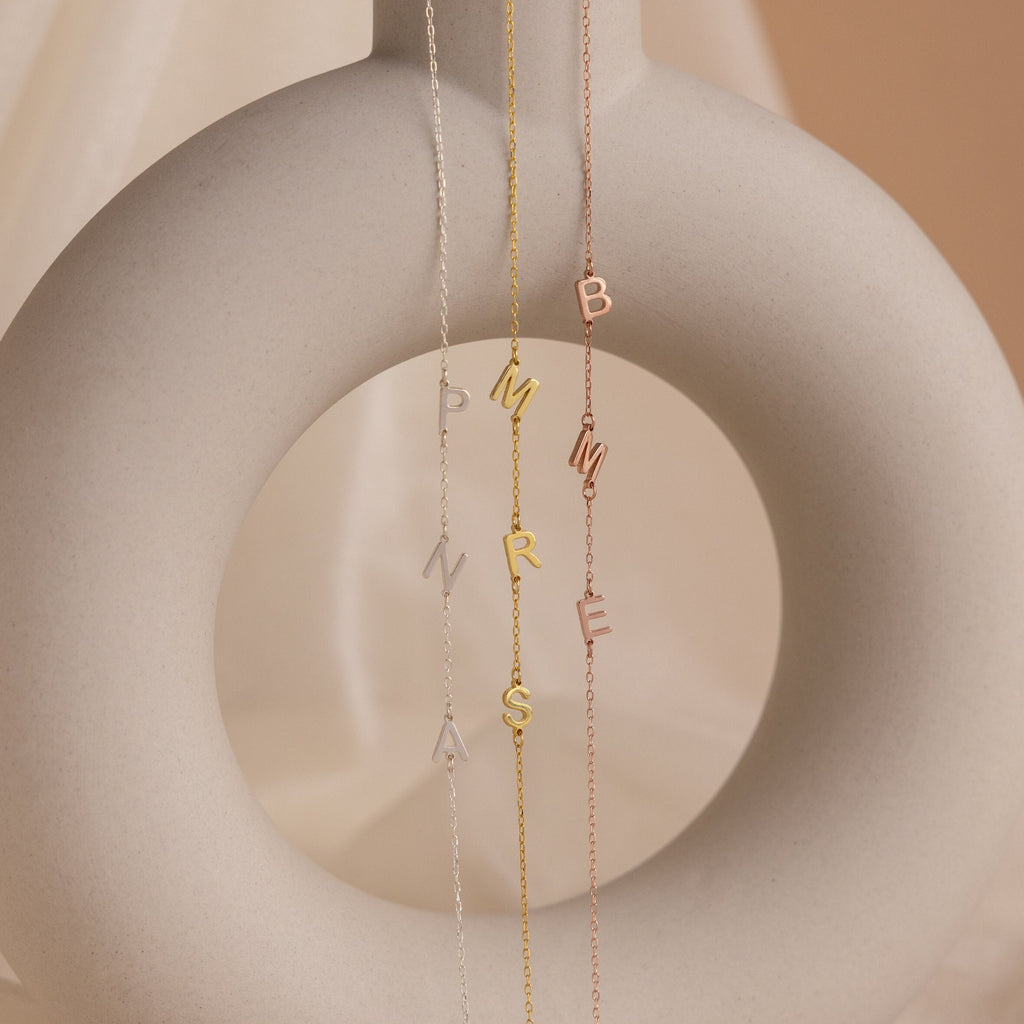 The Sideways Initial Necklace, featuring three delicate chain necklaces with letter charms, drapes elegantly on a light beige ceramic sculpture—making it a thoughtful and unique Christmas gift.