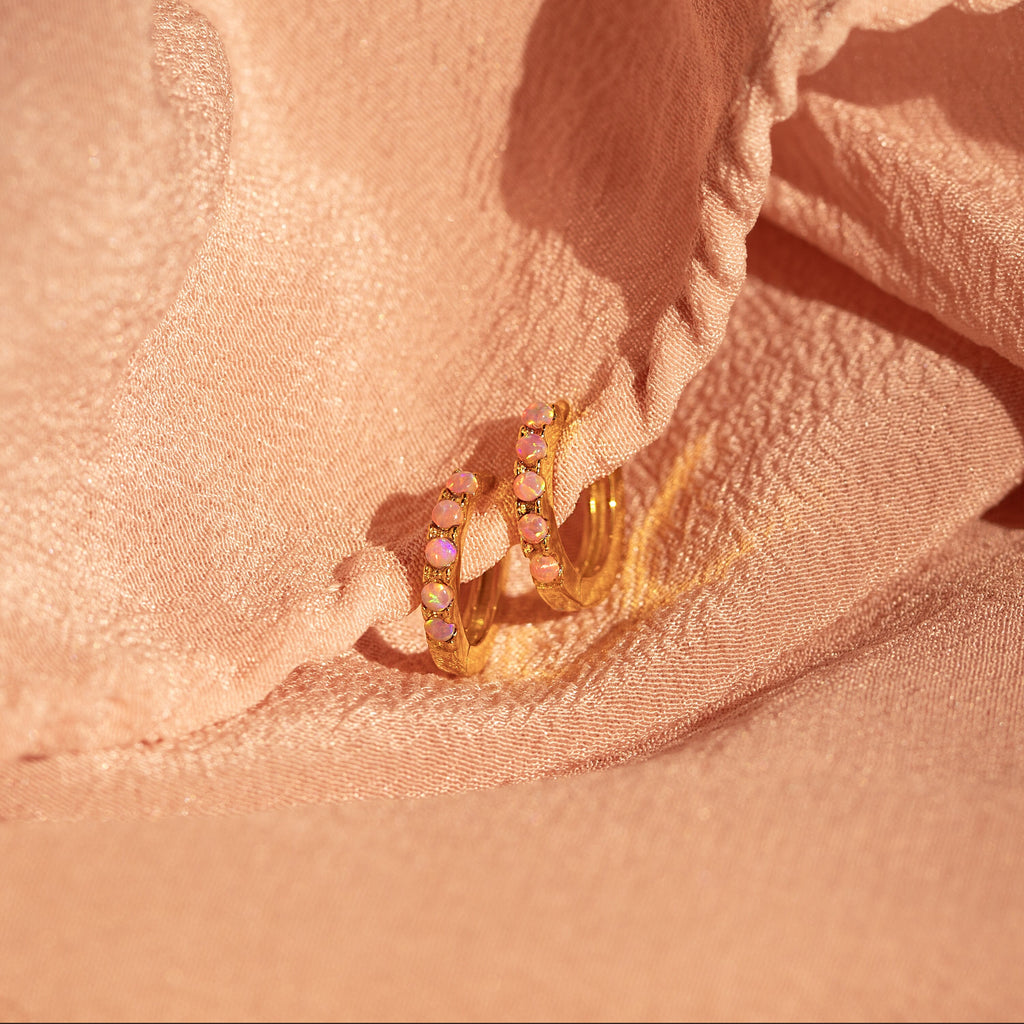 Pink Opal Huggies, elegant gold hoop earrings with pastel gemstones, are displayed on soft peach-colored fabric for a chic, feminine jewelry showcase.