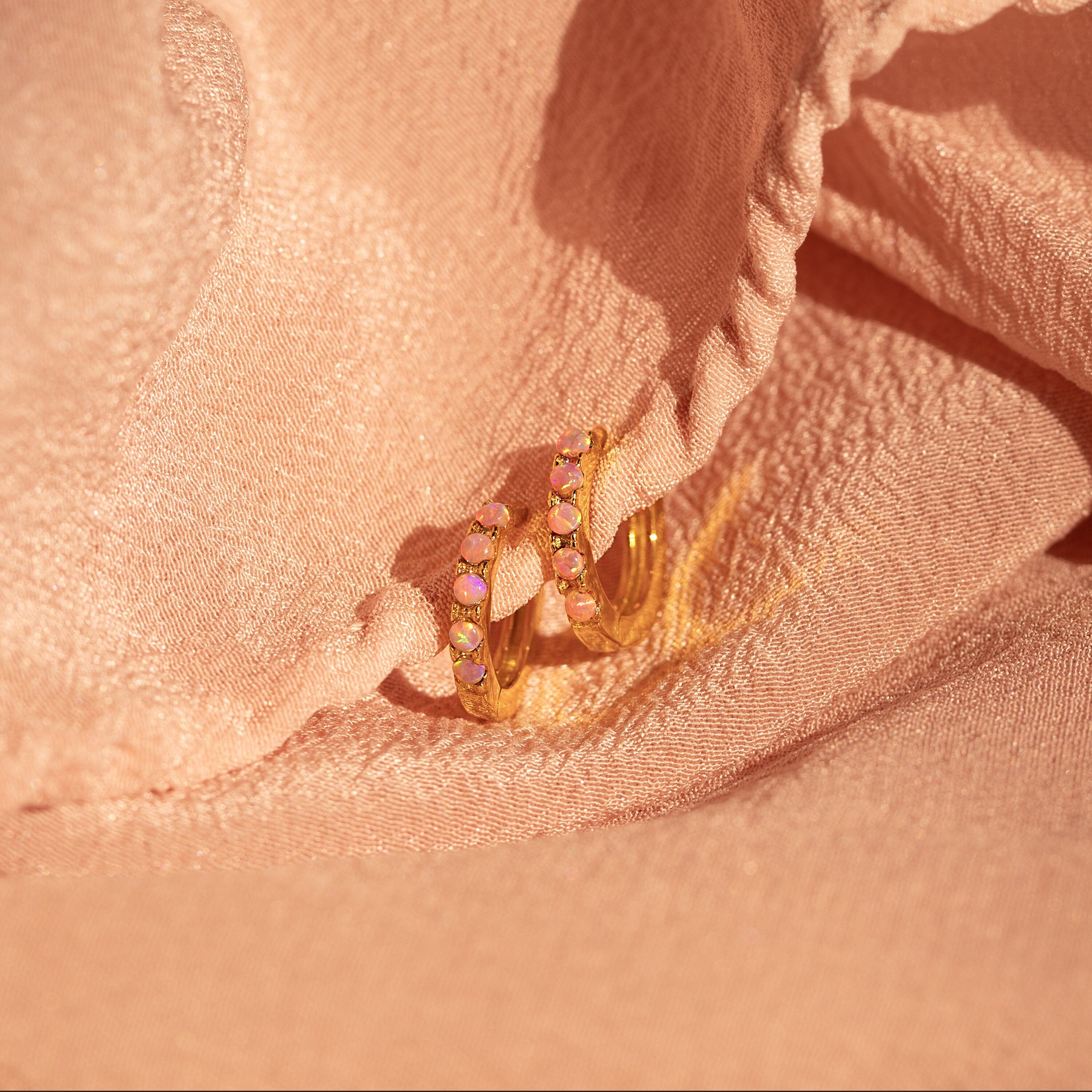 Pink Opal Huggies, elegant gold hoop earrings with pastel gemstones, are displayed on soft peach-colored fabric for a chic, feminine jewelry showcase.