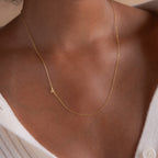 The Venice Sideways Initial Necklace, featuring a gold "A" pendant, rests elegantly on a person's neck above a white ribbed shirt, showcasing stylish personalized jewelry.