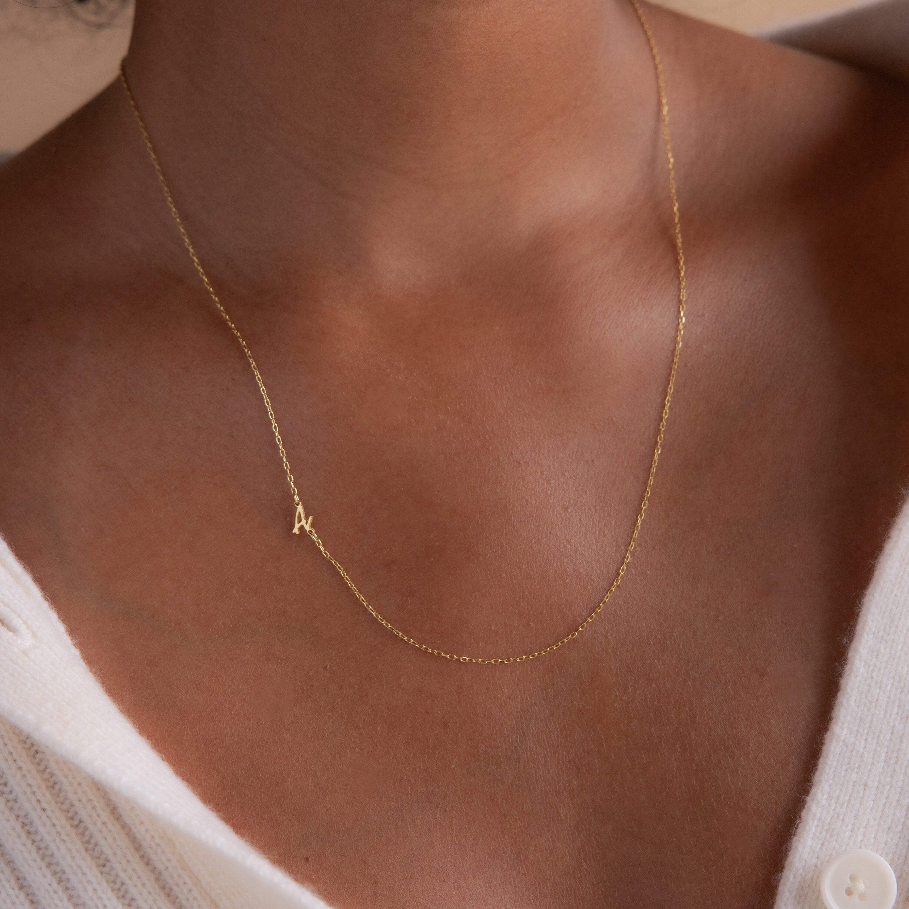 The Venice Sideways Initial Necklace, featuring a gold "A" pendant, rests elegantly on a person's neck above a white ribbed shirt, showcasing stylish personalized jewelry.
