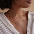 A woman wearing a cream-colored ribbed buttoned sweater and the Sideways Initial Necklace In Box Chain, layered with other gold necklaces.