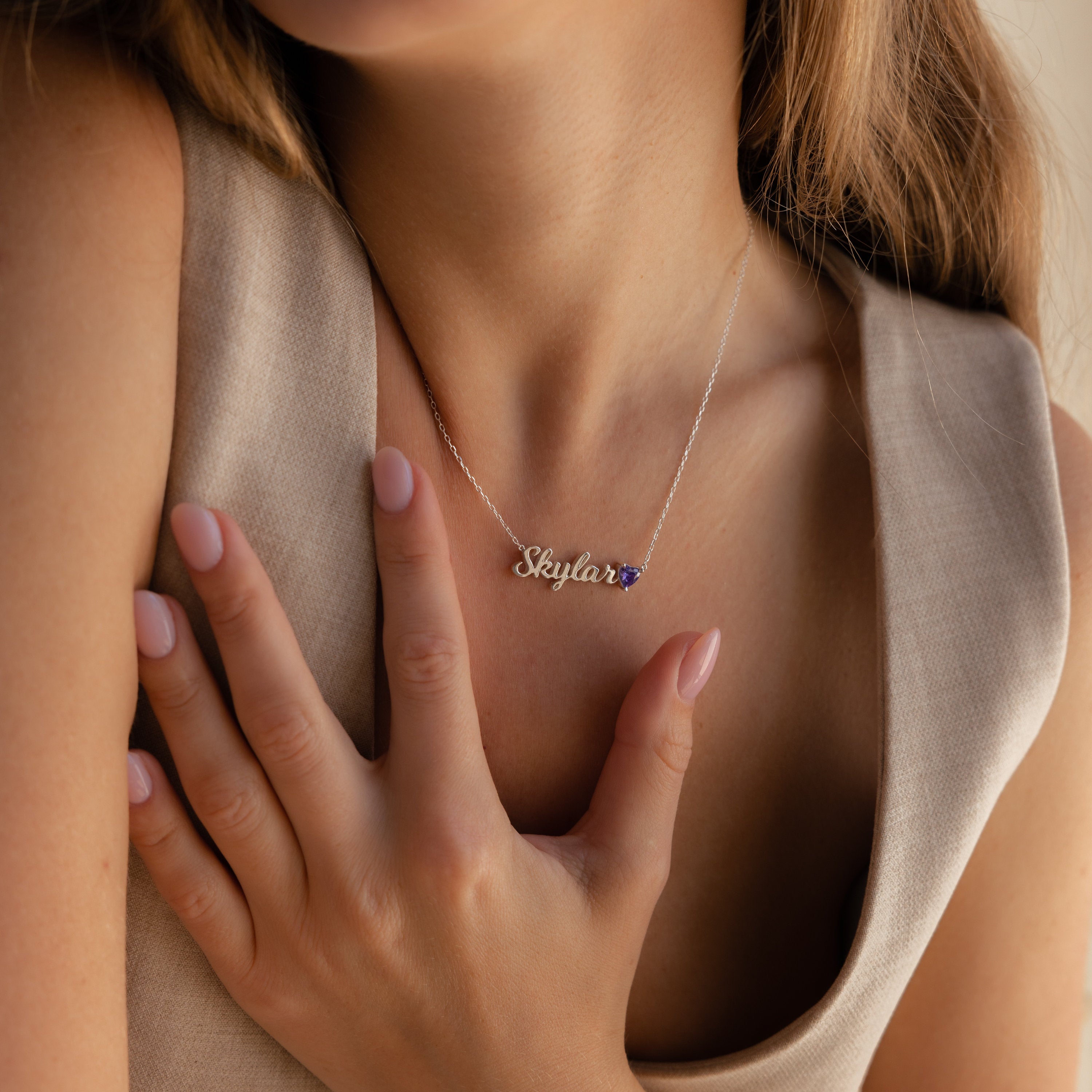 A woman wearing a minimalist Mellow Name Heart Birthstone Necklace with "Skylar," touches her chest with manicured fingers.