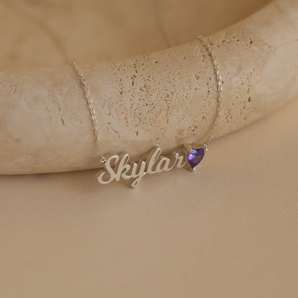 The Mellow Name Heart Birthstone Necklace, personalized with "Skylar" and a small purple heart charm, is a minimalist jewelry piece shown on a beige surface.