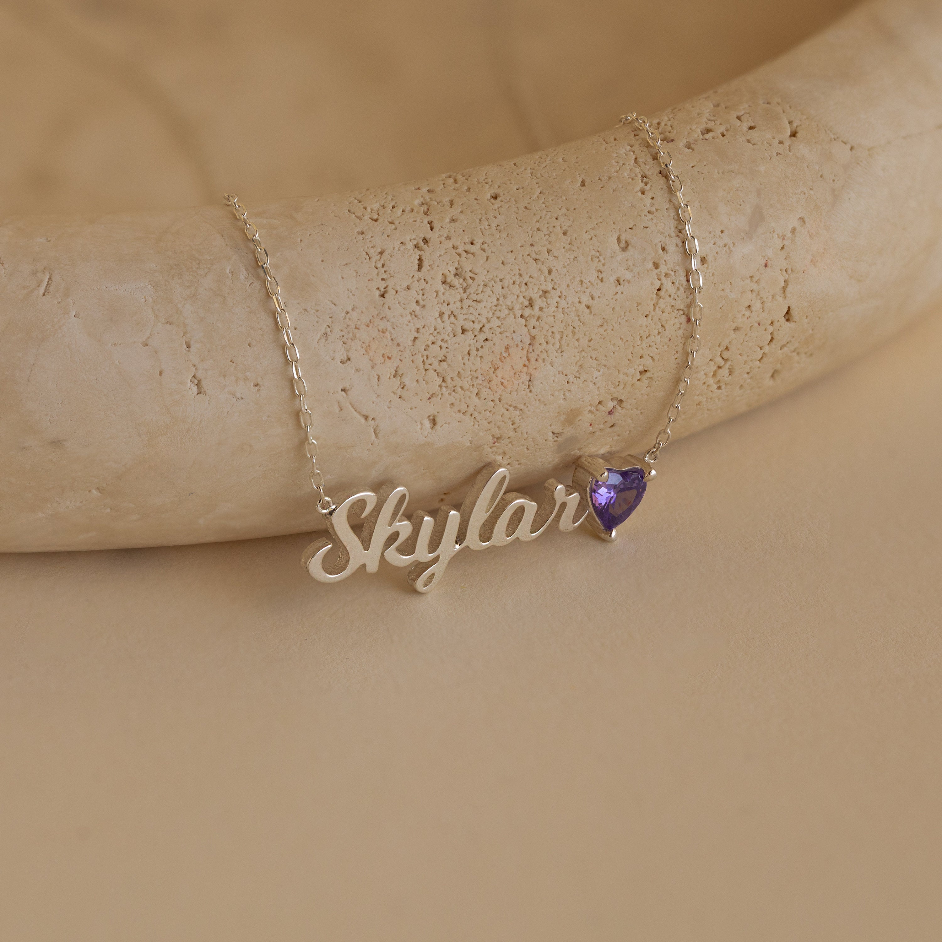 The Mellow Name Heart Birthstone Necklace, personalized with "Skylar" and a small purple heart charm, is a minimalist jewelry piece shown on a beige surface.