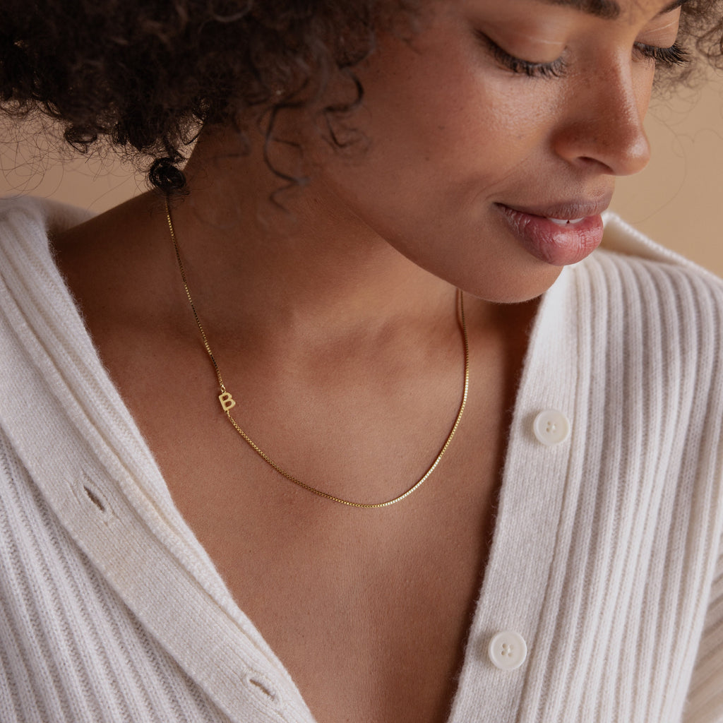 Woman wearing a white ribbed top and the Sideways Initial Necklace In Box Chain with a small gold B pendant, looking down and smiling, showcasing chic personalized jewelry.