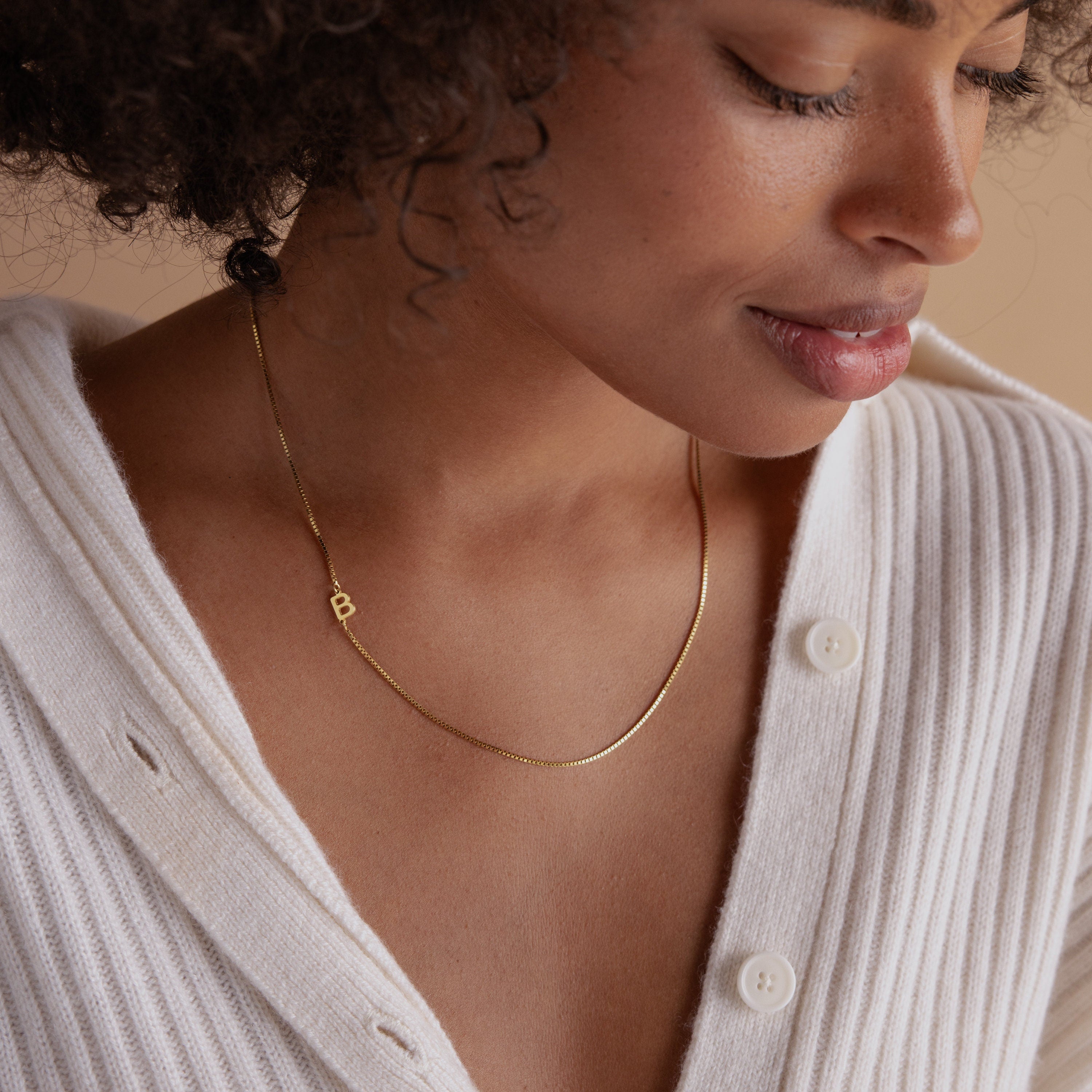 Woman wearing a white ribbed top and the Sideways Initial Necklace In Box Chain with a small gold B pendant, looking down and smiling, showcasing chic personalized jewelry.