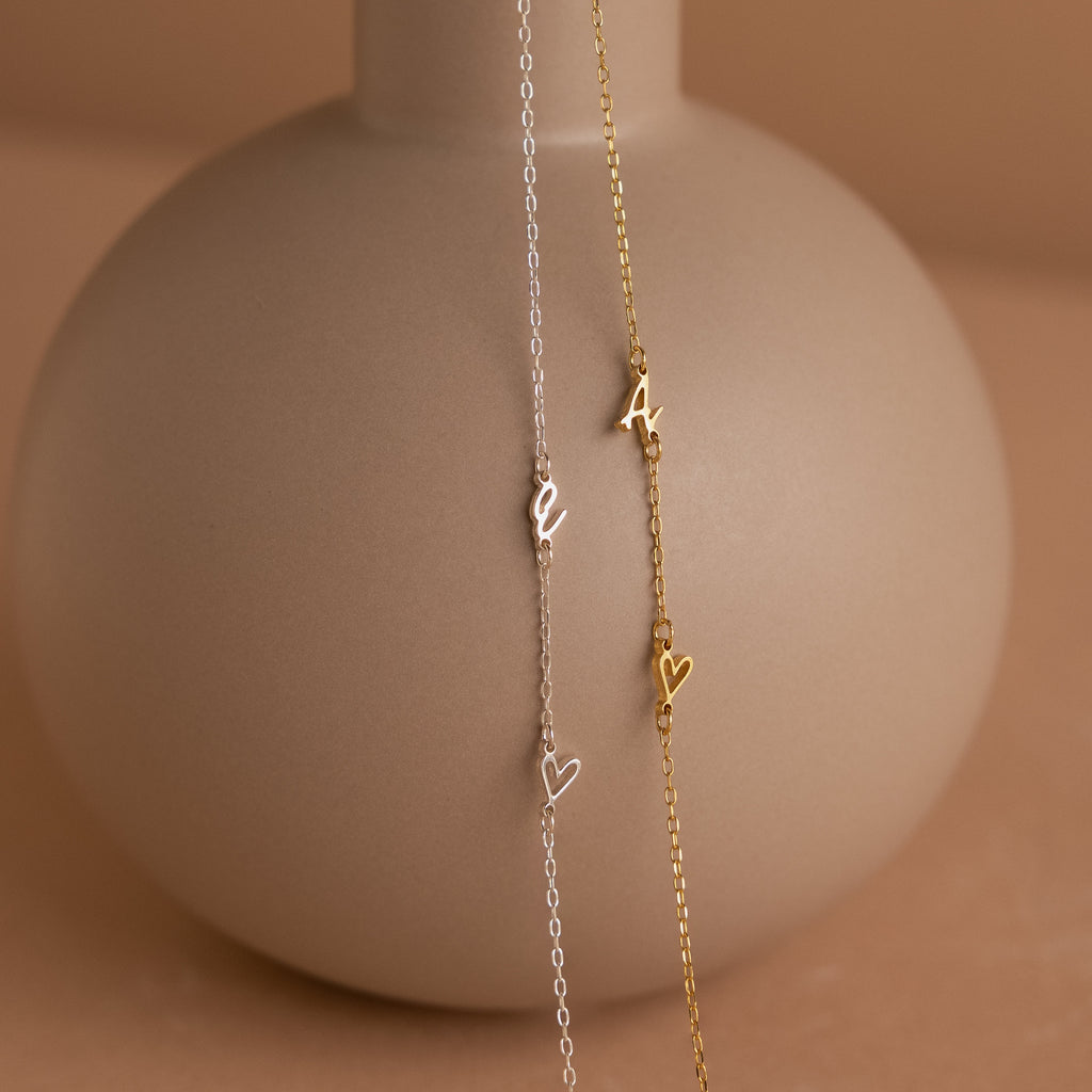 The Sideways Initial Heart Necklace pairs perfectly with delicate charm bracelets—styled here with heart and letter charms on a matte beige vase.