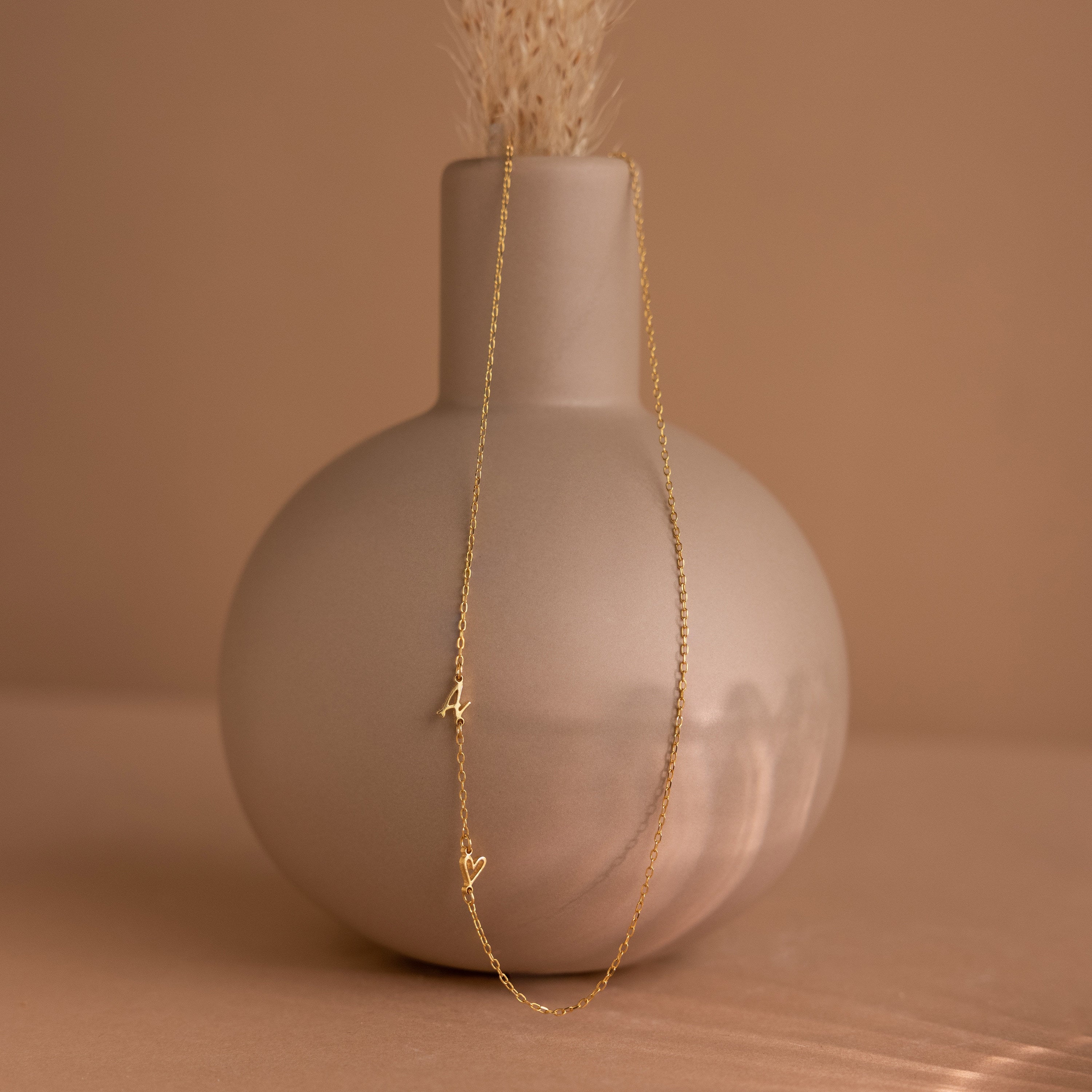 Sideways Initial Heart Necklace featuring a gold chain with a heart pendant and letter A, displayed draped over a matte beige vase on a neutral background.