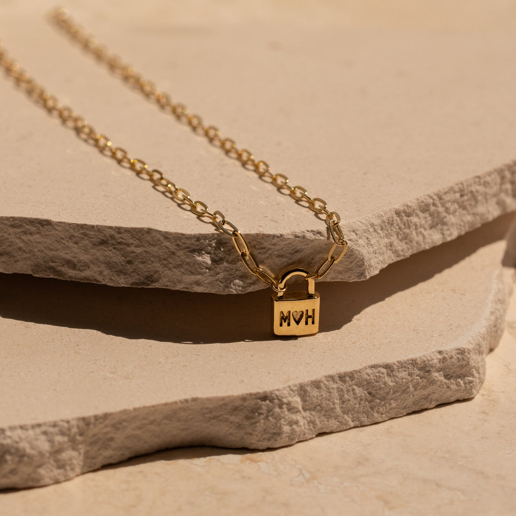 The Initial Padlock Paperclip Necklace features a small engraved pendant, displayed on textured stone slabs—a stylish personalized accessory, perfect for couples or as a unique gift.