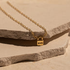 The Initial Padlock Paperclip Necklace features a small engraved pendant, displayed on textured stone slabs—a stylish personalized accessory, perfect for couples or as a unique gift.