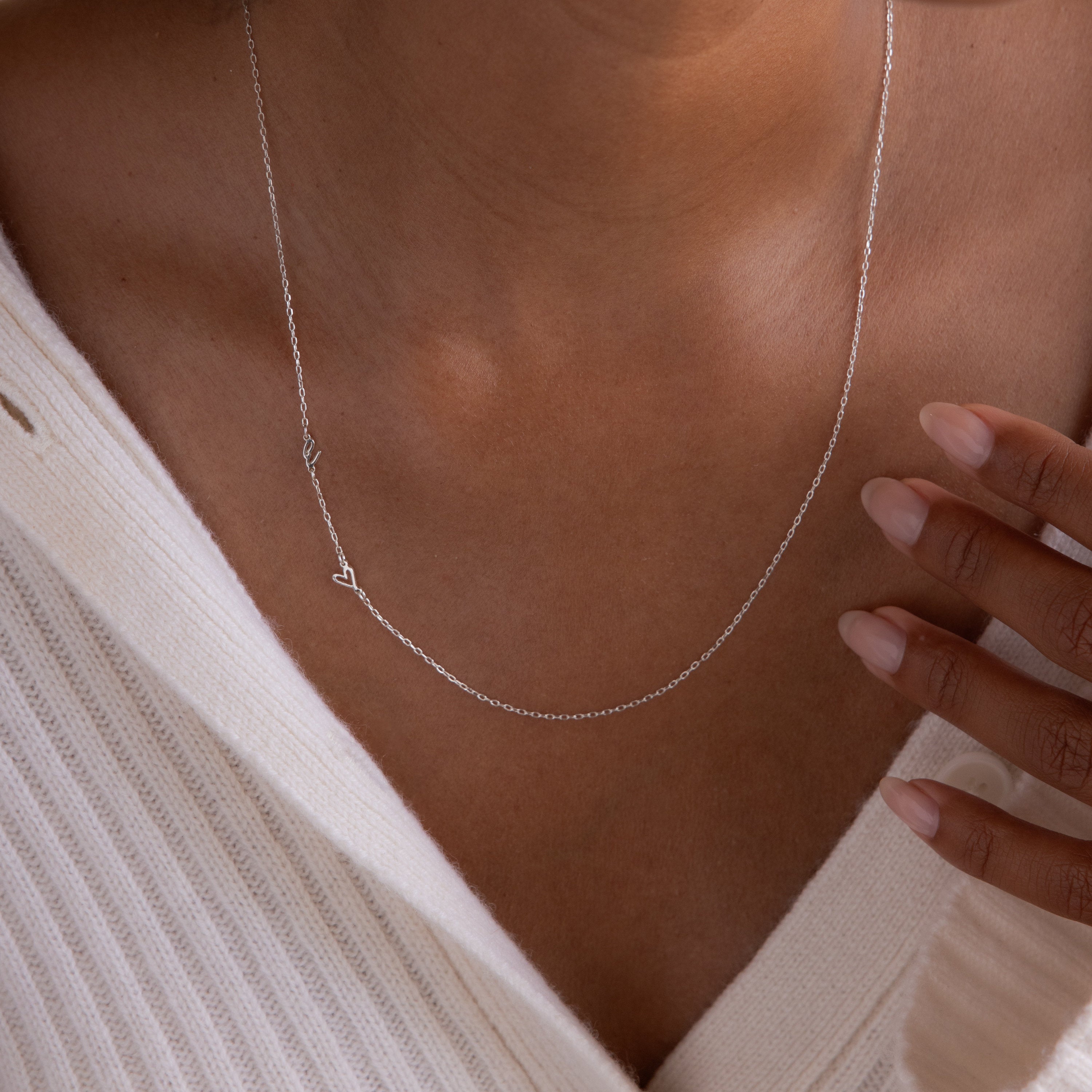 A person in a cream ribbed sweater displays manicured fingers near the collar, highlighting the elegant Sideways Initial Heart Necklace.