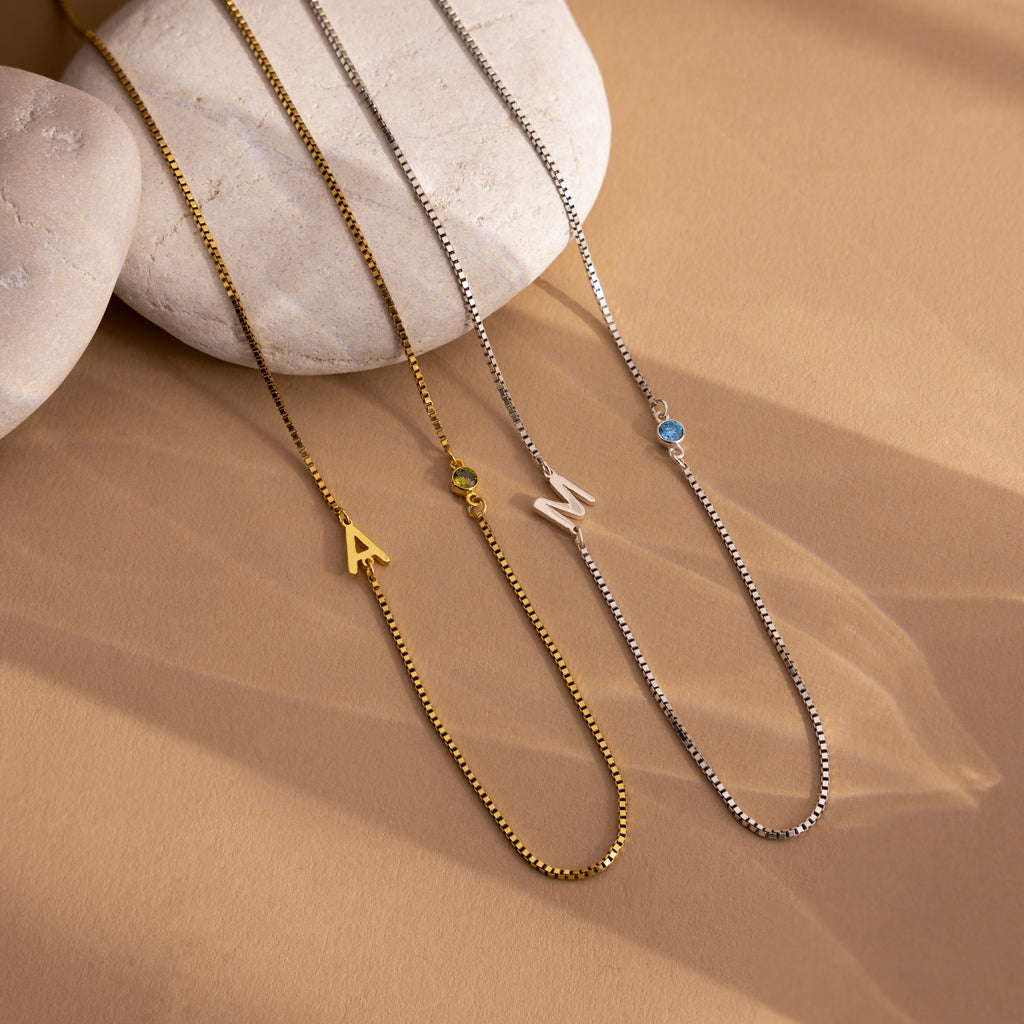 Two Birthstone Initial Necklaces in Box Chain, featuring “A” and “M” letter pendants each with a small gemstone, are displayed on beige stones for a personalized and timeless look.