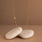 The Birthstone Initial Necklace in Box Chain features a small A pendant with a green gem, displayed on smooth beige stones. Its elegant box chain and personalized initial make this piece charming and unique.
