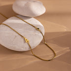 The Birthstone Initial Necklace in Box Chain rests on smooth white stones atop a beige surface—a perfect personalized accessory for any occasion.