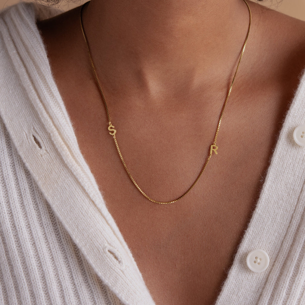 A woman wears the Duo Initial Necklace in Box Chain featuring S and R letter charms over a white ribbed sweater.