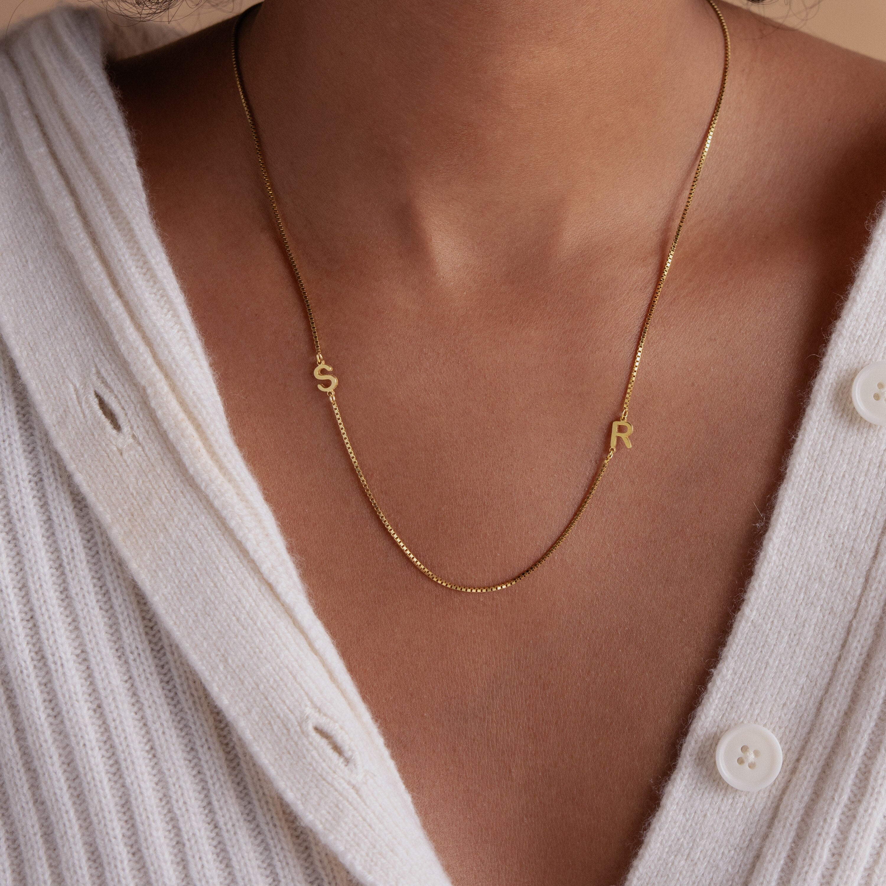 A woman wears the Duo Initial Necklace in Box Chain featuring S and R letter charms over a white ribbed sweater.