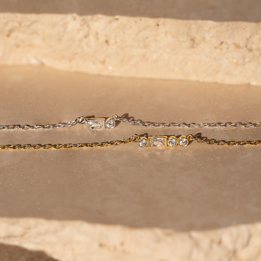 Gold and silver chain bracelets with rectangular and round gemstones rest on a stone surface in sunlight—perfect for pairing with a personalized jewelry piece like the Initial Morse Code Necklace.