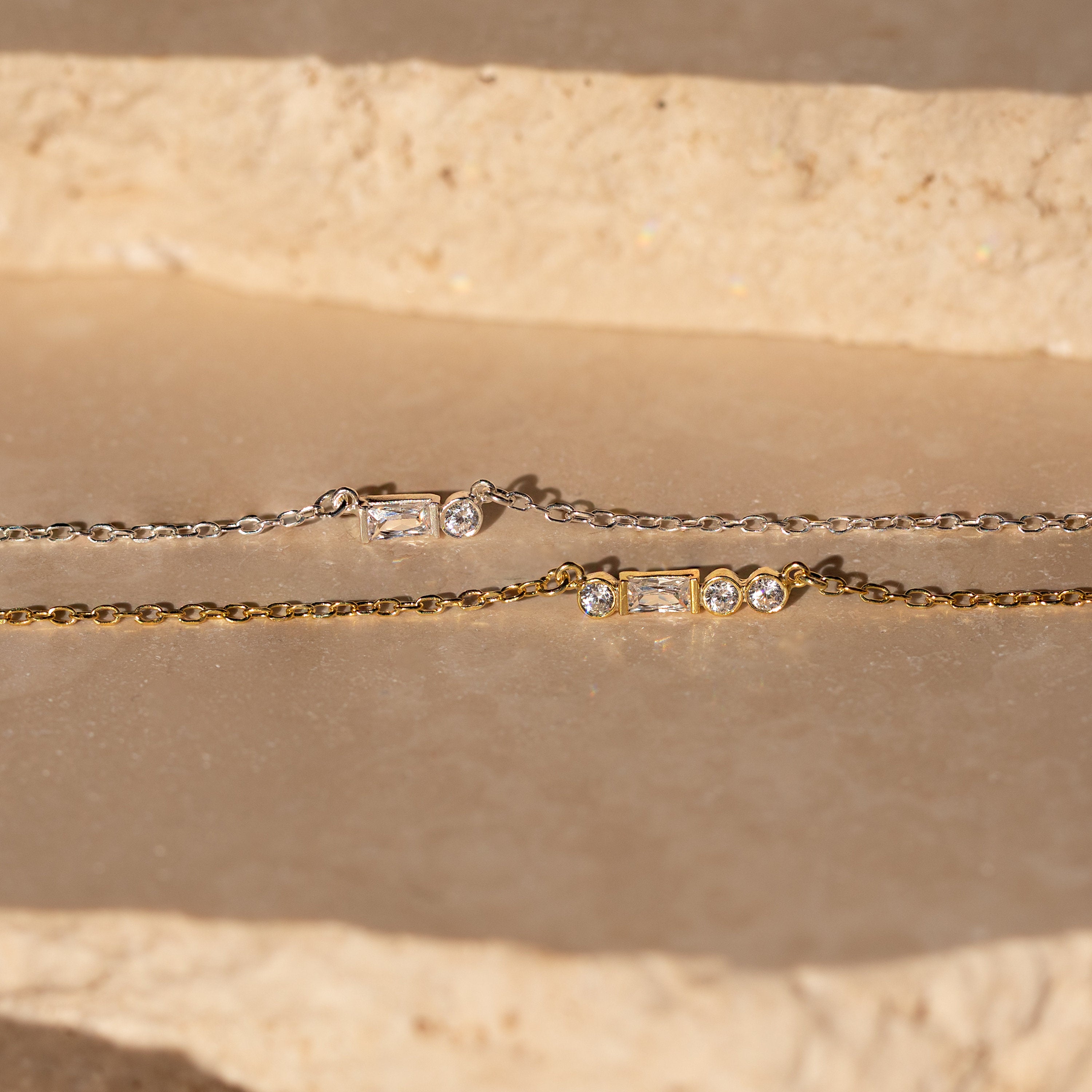 Gold and silver chain bracelets with rectangular and round gemstones rest on a stone surface in sunlight—perfect for pairing with a personalized jewelry piece like the Initial Morse Code Necklace.
