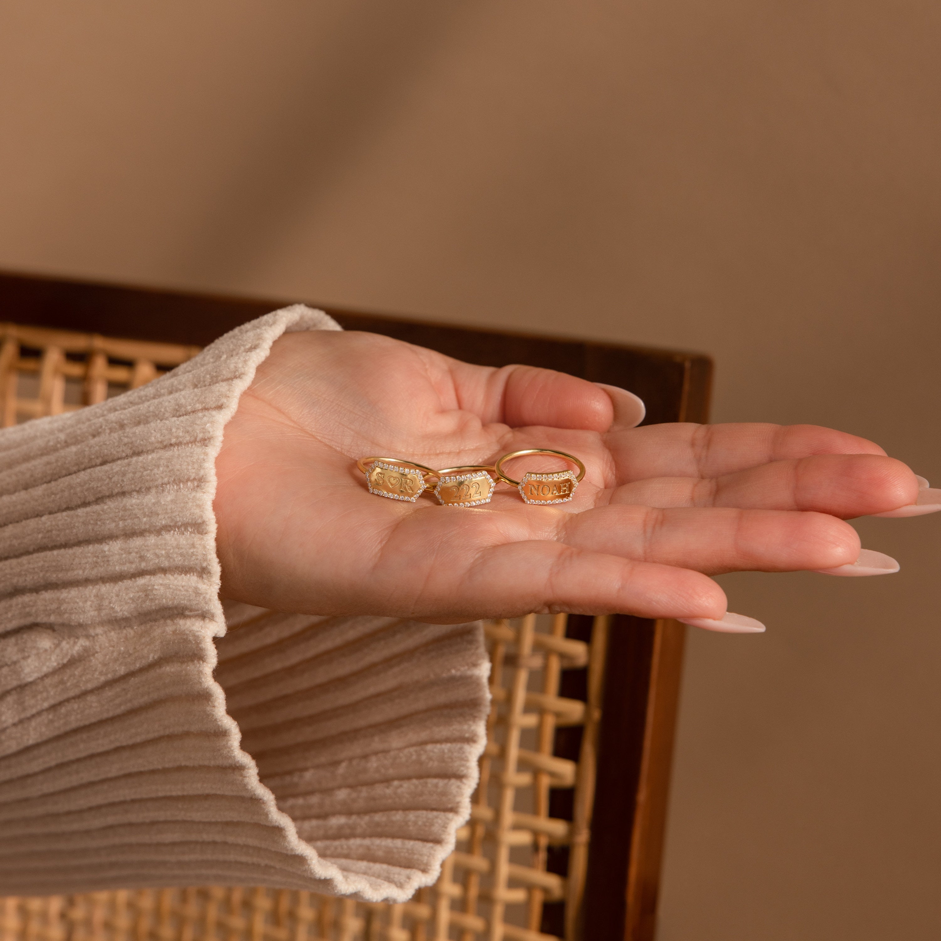A hand in a beige sleeve holds three gold rings, featuring the Custom Pave Hexagon Ring, above a woven chair with a brown background.