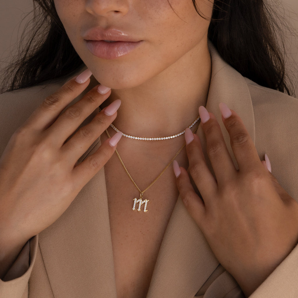A woman wears a beige blazer and layered gold necklaces, including the Gothic Pave Initial Necklace, while showing manicured nails painted light pink.