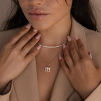 A woman wears a beige blazer and layered gold necklaces, including the Gothic Pave Initial Necklace, while showing manicured nails painted light pink.