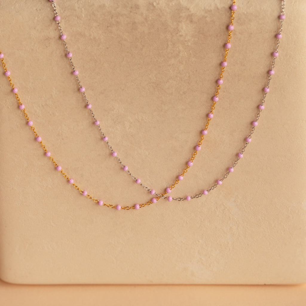 The Pink Station Necklace, a delicate beaded pink necklace, is showcased on a beige textured background—ideal for layering with other necklaces.
