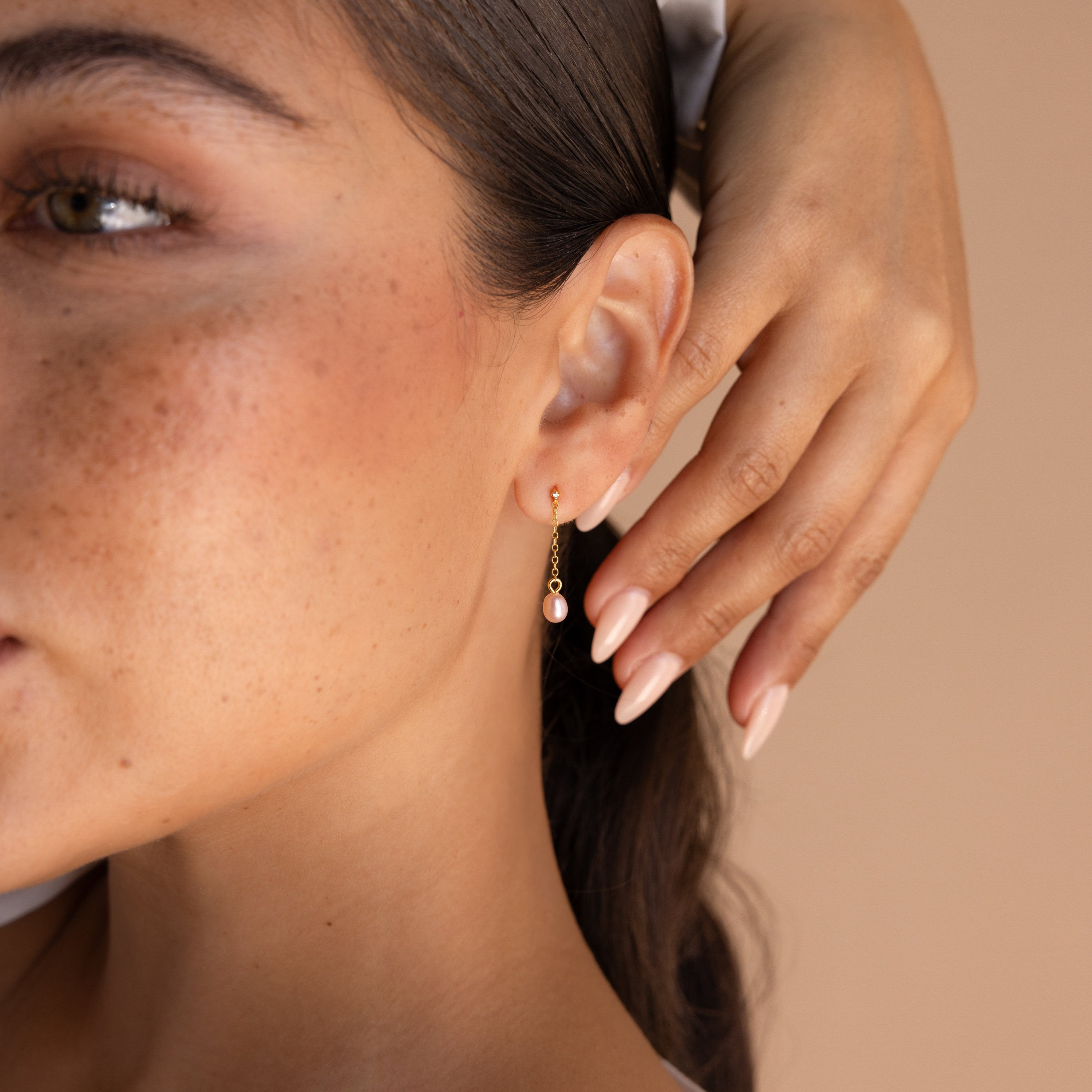 A woman with freckled skin touches her ear, wearing the elegant Isha Pink Pearl Earrings featuring a gold drop design with a peach-colored stone.