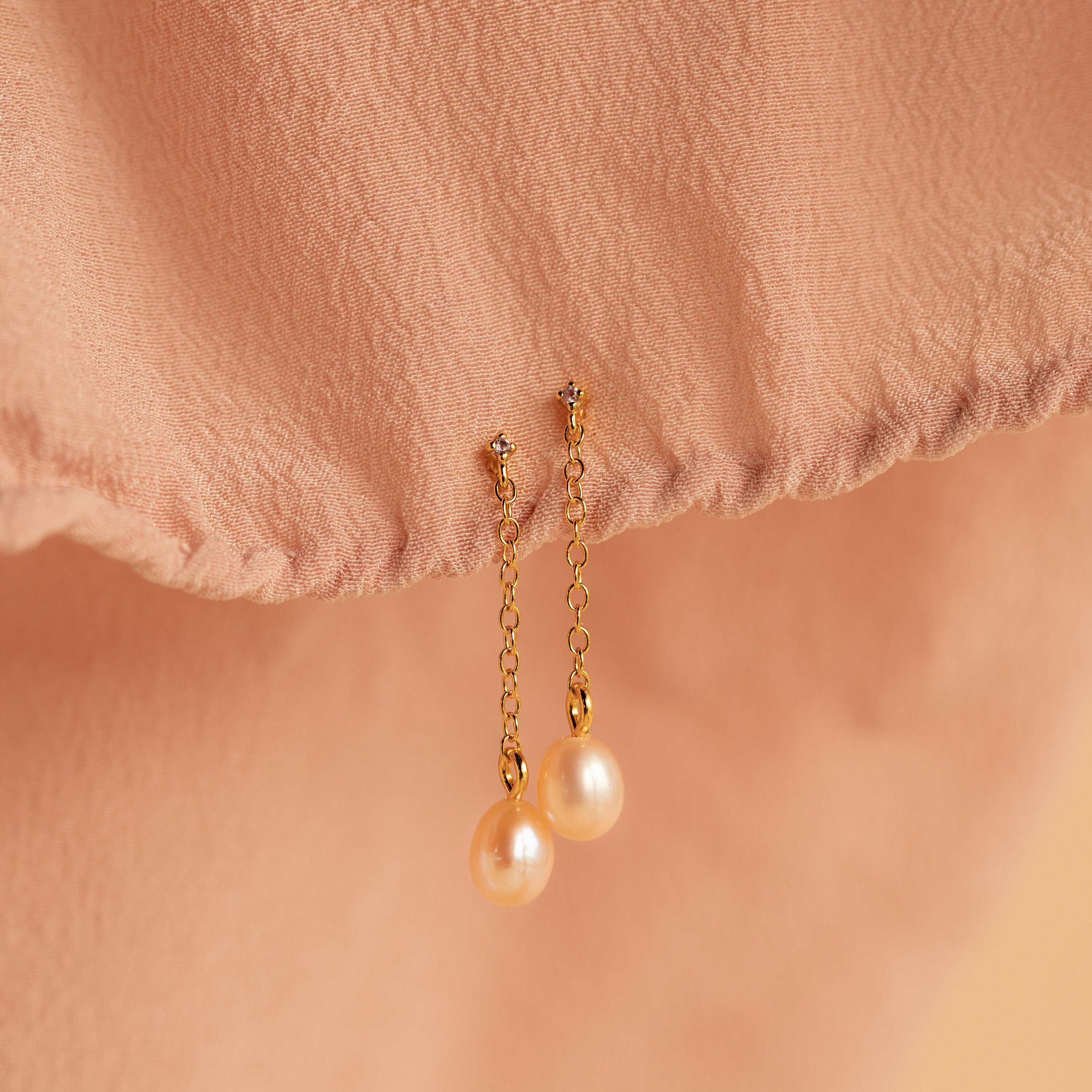 The Isha Pink Pearl Earrings, featuring gold chains and pearl drops against a peach fabric background, add an elegant touch of sophistication to any look.