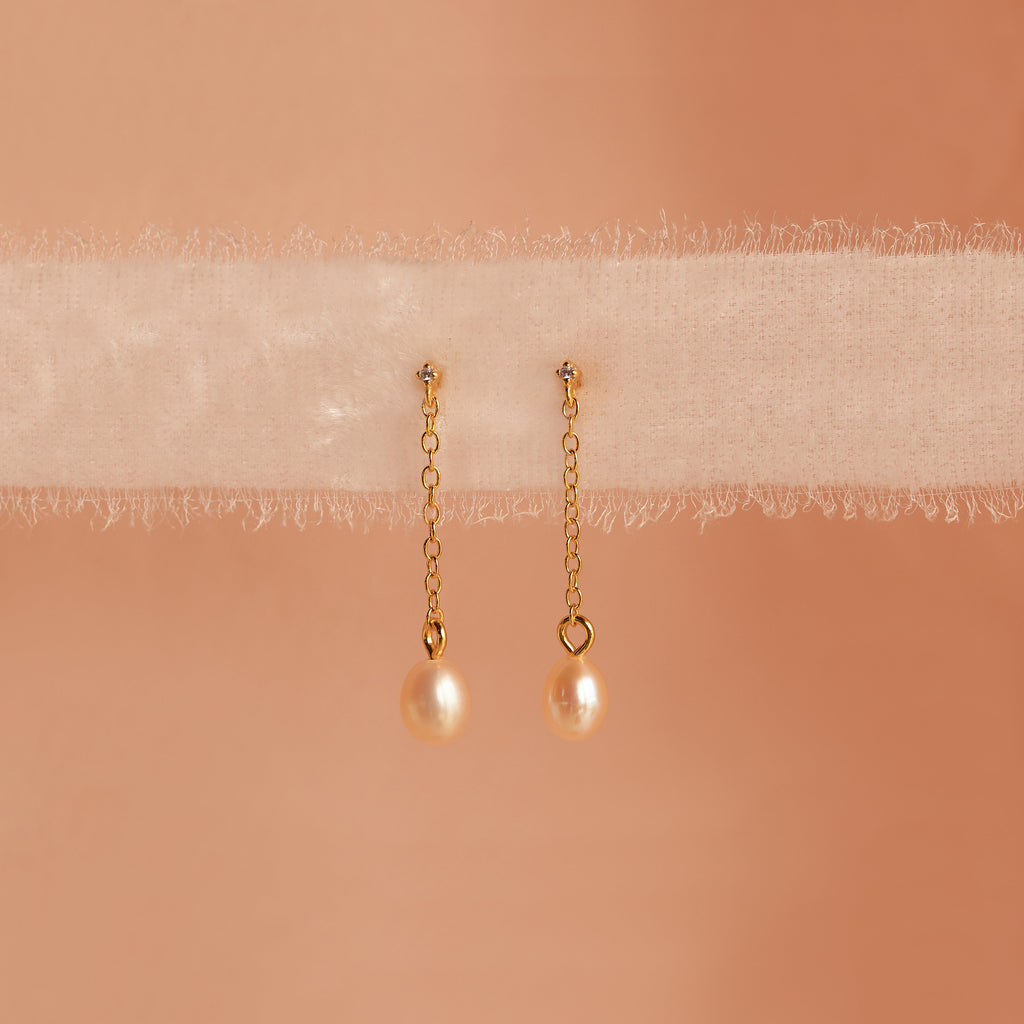 Isha Pink Pearl Earrings feature gold chains with pearl drops, elegantly displayed on soft, sheer fabric against a peach background—showcasing the timeless charm of these delicate dangling earrings.