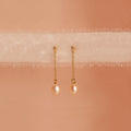 Isha Pink Pearl Earrings feature gold chains with pearl drops, elegantly displayed on soft, sheer fabric against a peach background—showcasing the timeless charm of these delicate dangling earrings.