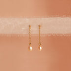 Isha Pink Pearl Earrings feature gold chains with pearl drops, elegantly displayed on soft, sheer fabric against a peach background—showcasing the timeless charm of these delicate dangling earrings.